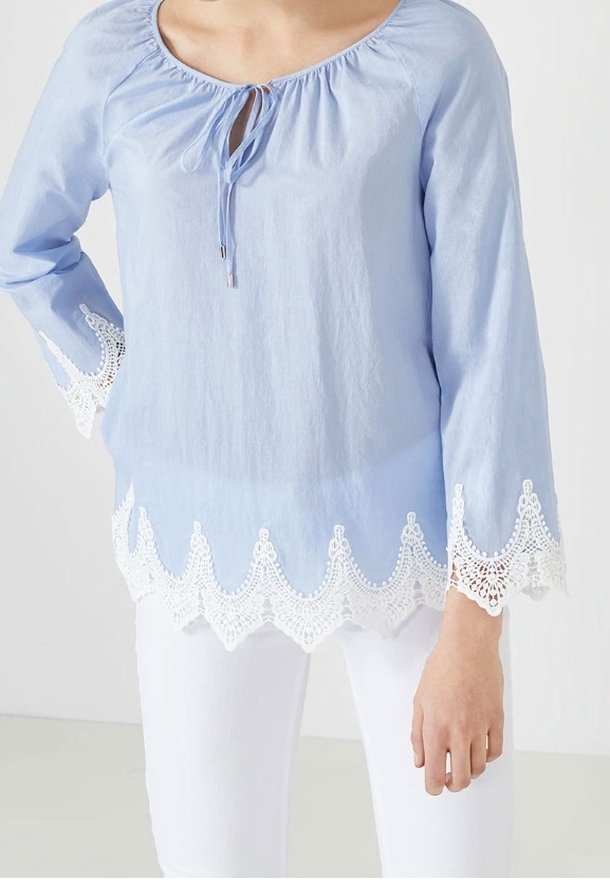 Ljusblå blus med långa ärmar, rund halsringning, knytdetalj fram, och vit spets- nederdel med vågiga kanter. Slät tygtextur.