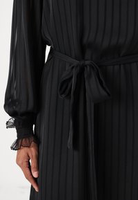 Črna črtasta obleka z pasom. Prosojna tkanina, dolgi rokavi z nabranimi manšetami. Gladka tekstura z diskretnim sijajem.