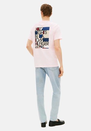 Hombre con camiseta rosa claro con estampado multicolor "Jazz Nights in L.A. Tommy Hilfiger est. 1985" en la parte trasera, vaqueros azul claro y zapatos negros.