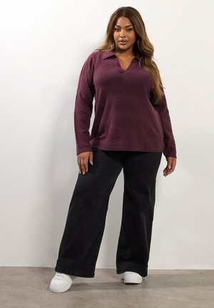 Jersey de lana púrpura de gran tamaño con escote en V y cuello, combinado con pantalones anchos negros y zapatillas blancas sobre un fondo neutral.