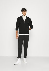 Felpa nera con zip e coulisse, indossata sopra un maglione bianco a collo alto. Abbinata a pantaloni scuri e sneakers bianche alte. Texture liscia.