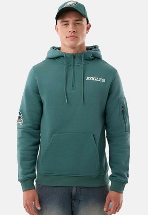 Uomo che indossa una felpa Eagles verde acqua con tasca anteriore, mezza cerniera e cappellino abbinato Eagles, in piedi davanti a uno sfondo semplice.