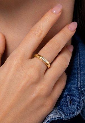 Anillo de oro con una banda lisa y una fila de pequeñas piedras transparentes colocadas en ángulo, llevado en una mano con uñas bien cuidadas.