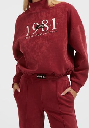 Kobieta ubrana w bordową bluzę Guess i dopasowane spodnie z białym napisem "1981" i "Unique Sporty Attitude", ręce w kieszeniach.