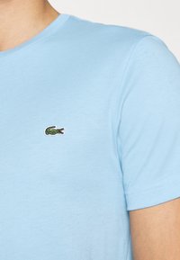 Světle modré bavlněné tričko s kulatým výstřihem. Na levé straně hrudi se nachází malá vyšitá zelená logo krokodýla. Měkký materiál, krátké rukávy.