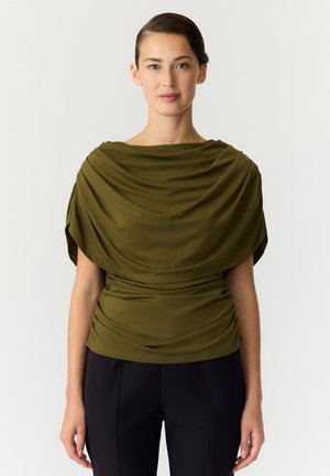 Femme portant un haut drapé vert olive et un pantalon noir, debout devant un fond clair uni, regardant droit devant.