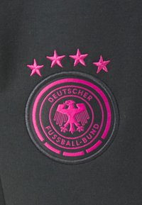 Zwarte stof met een geborduurd rond logo dat een roze adelaar toont, de tekst "DEUTSCHER FUSSBALL-BUND," en vier roze sterren boven het logo.