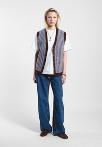 Gilet en tricot violet et marron à rayures verticales, avec un col en V, des boutons sur le devant, porté sur un t-shirt blanc et un jean bleu.