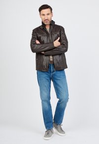 Bruine leren jas met hoge kraag, voorzakken en rits, gecombineerd met blauwe jeans en grijze sneakers, tegen een effen achtergrond.