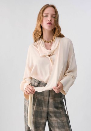 Femme aux longs cheveux ondulés portant un chemisier crème avec un détail foulard, un pantalon taille haute à carreaux et un collier épais en chaîne dorée.