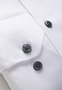 Valge triiksärgi mansett kahe tumehalliga nupuga, kus on nähtav riide detailne tekstuur ja õmblused.