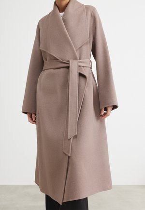 Kvinde iført lang taupe uldfrakke med brede revers og bindebælte, stående mod hvid baggrund.