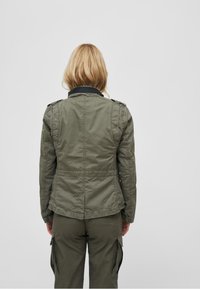 Olive green, fitted jacket with a zip front, textured fabric, epaulets on shoulders, and a contrasting black collar.