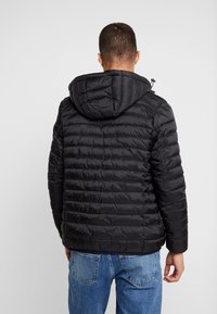 Schwarze isolierte Jacke mit horizontalem Steppmuster, ausgestattet mit Kapuze und Reißverschlüssen, kombiniert mit hellblauen Jeans.