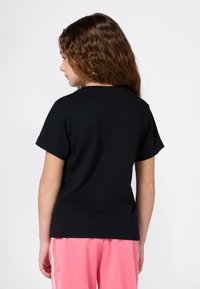 Chica con cabello castaño rizado que lleva una camiseta negra de mangas cortas y pantalones rosas, de lado contra un fondo blanco.