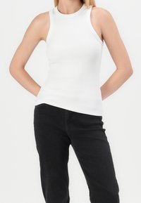 Donna che indossa un top bianco senza maniche a coste e pantaloni neri, in piedi con le mani dietro la schiena su uno sfondo bianco.