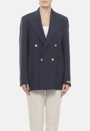 Persona che indossa una giacca doppiopetto blu navy con bottoni beige e grandi tasche frontali sopra una maglietta bianca e pantaloni beige.