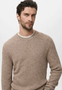 Man met kort donker haar en lichte baard die een beige gebreid sweater over een wit shirt draagt, recht naar voren kijkend tegen een effen lichte achtergrond.