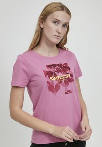 Lyserød bomulds T-shirt med korte ærmer, der har et blomstermotiv og teksten "Simplicity Made Perfect" i gul.