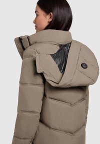 Beige pufferjack met een capuchon, met een zwarte voering. De stof is gestructureerd en doorgestikt, met een knoopaccent bij de nek.