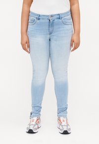 Šviesiai mėlynos aptemptos džinsus su išblukusia danga, priekinėmis kišenėmis ir mygtuko uždarymu. Derinami su pilkai oranžiniais sportiniais batais.