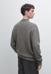 Pull en maille grise texturée avec un col rond et des poignets côtelés. Le design est décontracté, adapté à une tenue casual.