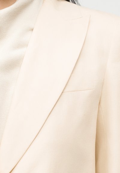 Nærbillede af en person iført en cremefarvet blazer over en tekstureret beige strik top, som viser jakkeens revers og brystlomme.