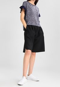 Koszulka w marynarskie wzory z falbaniastymi rękawami, w zestawieniu z czarnymi szortami. Model nosi białe sneakersy. Skupienie na swobodnym, luźnym kroju.