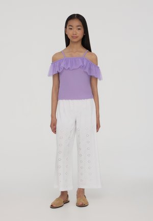 Lawendowa bluzka bez ramiączek z falbankami z tiulu, w zestawieniu z luźnymi białymi spodniami z wycięciami, oraz brązowymi butami na odkrytej pięcie.