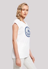 White cotton T-shirt with rolled sleeves, featuring a blue graphic design of a hand and waves. Model wears slim-fit white pants.