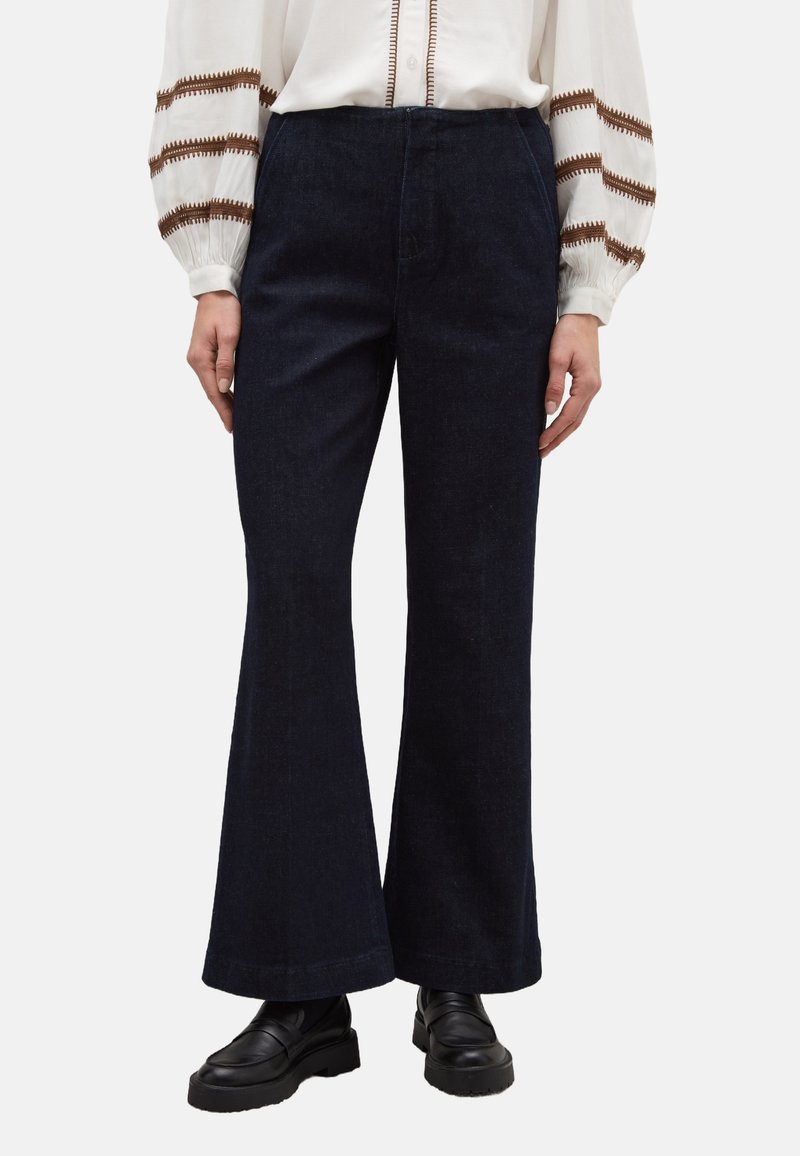 Dunkelblaue Jeans mit Schlag aus Denim, mit hoher Taille, zwei Vordertaschen und einem sauber verarbeiteten Saum. Kombiniert mit schwarzen Loafers.