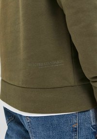 Olivově zelená mikina s ribovaným lemem a manžetami, vyrobená z měkké tkaniny. Na zádech se nachází logo, doplněná džínami.