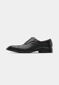 Zapato de vestir de cuero negro con textura suave, puntera redondeada y cinco ojales. Presenta un tacón bajo apilado y un sutil detalle de costura.