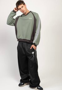 Felpa verde e grigia con scollo rotondo e dettagli scuri, abbinata a pantaloni neri larghi con logo bianco e strisce laterali.