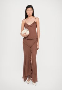 Brown, flowy dress with ruffled accents at the neckline and hem, featuring a side slit. Model holds a small, cream clutch purse.