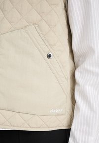 Beige quilted vest with diagonal pocket, silver snap button, and embroidered "Barbour" logo at the hem. Textured fabric detail visible.