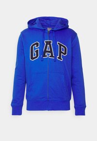 Hoodie bleu zippé avec un grand logo "GAP" blanc sur le devant, doté d'une capuche à cordon et de deux poches avant. Tissu doux et extensible.