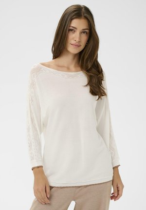 Witte, losvallende top met brede halslijn en mouwen met kantpatroon. Soepele textuur, afgewerkt met een gegolfde zoomdetail.