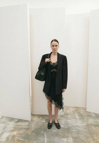 Blazer negro sobre un vestido negro con encaje en el borde y dobladillo asimétrico. La modelo sostiene un bolso negro. El suelo presenta un patrón texturizado beige y gris.