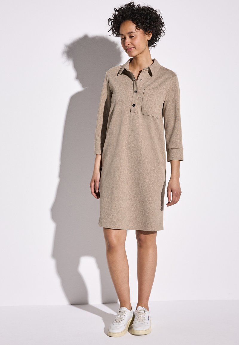 Beige jurk met een kraag met knopen, driekwart mouwen en een borstzak. Zachte stof en rechte silhouet, gecombineerd met witte sneakers.