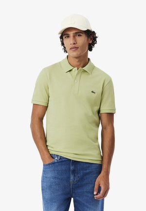 Uomo che indossa una polo verde chiaro, jeans blu e cappello beige, in piedi con una mano in tasca contro uno sfondo bianco.