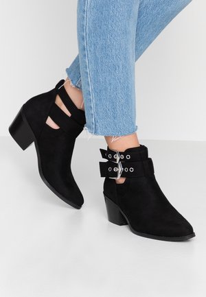 Schwarze Wildleder-Stiefeletten mit doppelten Schnallenriemen und Blockabsätzen, getragen zu hellblauen, kurz geschnittenen Jeans mit ausgefranstem Saum auf weißem Hintergrund.