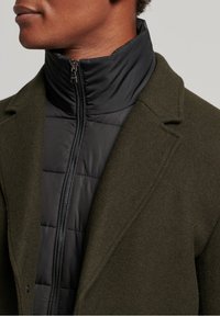 Manteau vert olive avec un col montant par-dessus une doudoune noire zippée. Le manteau présente un tissu texturé et un design structuré.