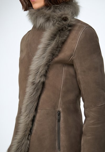 Manteau en daim marron avec un col en fourrure grise, des poches latérales zippées et une coupe ajustée. Le tissu est lisse avec une texture douce.
