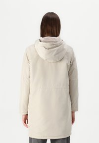 Lys beige parkajakke med hette, med et mykt innerfôr, lange ermer og innsvingt midje. Glatt tekstur og knelength design.