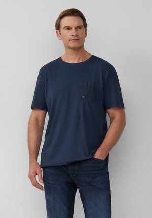 Marineblaue Baumwoll-T-Shirt mit rundem Halsausschnitt, kurzen Ärmeln und einer Brusttasche mit einem Patch-Logodetail. Getragen mit einer dunklen Jeans.