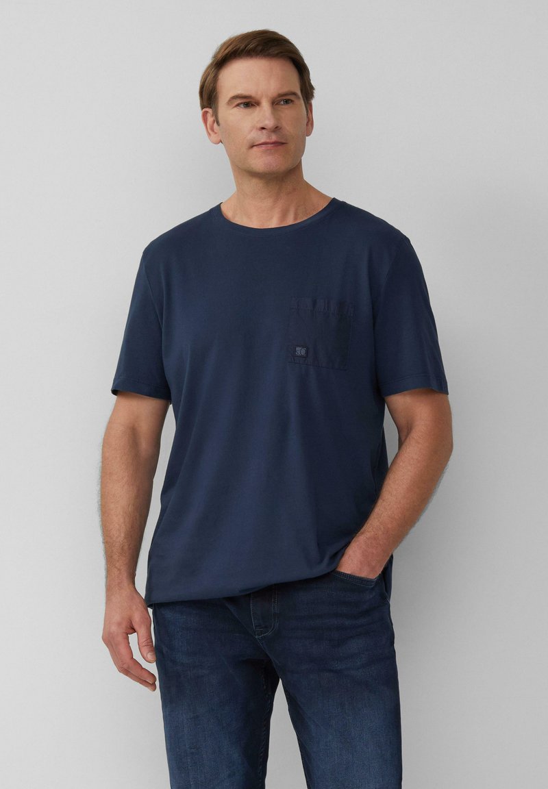 Navyblauwe katoenen t-shirt met een ronde hals, korte mouwen en een borstzak met een patchlogo detail. G gedragen met donkere spijkerbroek.