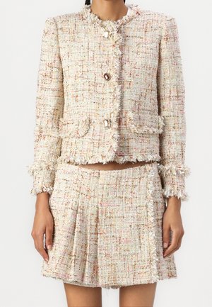 Femme portant une veste en tweed couleur crème et une jupe plissée assortie avec des bords effilochés et des fils multicolores.