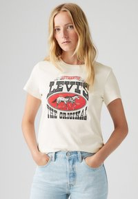 Kremkrāsas kokvilnas t-krekls ar retro Levi's grafiku, kurā attēloti zirgi, sarkans ovāls kontūrs un resna melna teksts. Valkāts ar gaiši ziliem džinsiem.