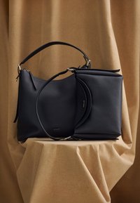 Fekete bőr kézitáska texturált felülettel, dupla fogantyúval, cipzáros záródással és aranyszínű kiegészítőkkel. Minimalista logó díszíti.